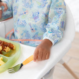 A child's hand, in a Bumkins Sleeved Bib: Aloha Stitch, holds a fork next to eggs and waffles on a sectioned plate in a high chair.