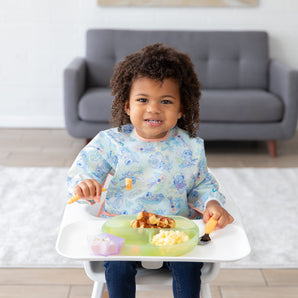A toddler with curly hair smiles in a high chair, enjoying breakfast on a green plate, wearing a Bumkins Sleeved Bib: Aloha Stitch.