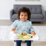 A toddler with curly hair smiles in a high chair, enjoying breakfast on a green plate, wearing a Bumkins Sleeved Bib: Aloha Stitch.