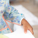 A child's arm in a Bumkins Sleeved Bib: Aloha Stitch rests on a high chair's white armrest.