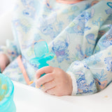 A child in a Bumkins Sleeved Bib: Aloha Stitch, holding a spoon, sitting near a bowl on a high chair tray.