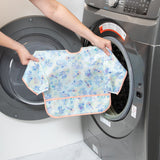 A person holds a Bumkins Sleeved Bib: Aloha Stitch in front of a front-loading washing machine.