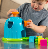 A child engages with the Fat Brain Toy Cos Shape Factory, boosting spatial reasoning by fitting blocks into a blue and green container.