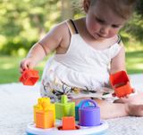 A toddler in a white dress enjoys enhancing fine motor skills with the colorful Plugzy Shape Learning Toy by Fat Brain Toy Co outdoors.