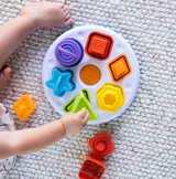 A child hones motor skills with the Plugzy Shape Learning Toy by Fat Brain Toy Co on a carpet, arranging colorful pieces like stars.