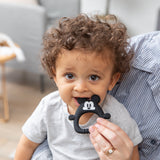 A curly-haired toddler chews a Bumkins Silicone Flat Teether: Mickey Mouse, while sitting on an adult's lap in a cozy room.