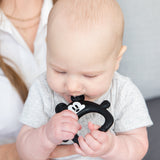 A bald baby in a gray shirt sits on a lap, eagerly chewing on a Bumkins Silicone Flat Teether: Mickey Mouse.