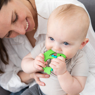 A woman smiling at a baby who is chewing on a green teething toy. The baby is wearing a light gray shirt and sitting on the womans lap. They are in a cozy indoor setting.