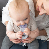 A baby joyfully chews a Bumkins Silicone Teether: Nintendo SNES™ Controller for ultimate gum relief on a woman's lap.