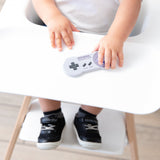A toddler grips a Bumkins Silicone Teether: Nintendo SNES™ Controller in a high chair with black sneakers and white socks visible.