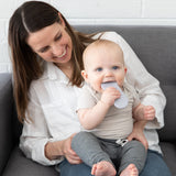 A woman smiles at a baby chewing a Bumkins Silicone Teether: Nintendo SNES™ Controller, with a white brick wall behind them on a gray couch.
