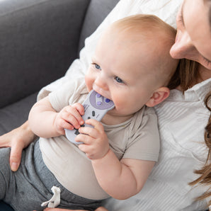 A baby chews a Bumkins Silicone Teether: Nintendo SNES™ Controller while sitting on an adult's lap on a gray couch.