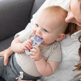 A baby chews a Bumkins Silicone Teether: Nintendo SNES™ Controller while sitting on an adult's lap on a gray couch.