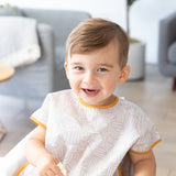 A happy toddler sits in a highchair wearing a Bumkins Junior Bib: Wander. A blurry living room with a couch and plant is in the background.
