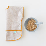 Bumkins Junior Bib: Wander with a catch-all pocket lays folded next to a bowl of cereal and baby spoon on a white background.