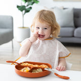 A blonde toddler in a Bumkins Junior Bib: Wander smiles while eating from a divided plate in a brightly lit room.