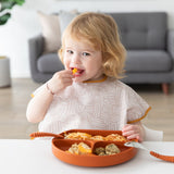 A blonde toddler eats from an orange plate at a white table, wearing a Bumkins Junior Bib: Wander. A gray sofa is in the background.