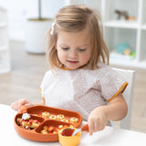 A toddler wears a Bumkins Junior Bib: Wander, while eating from a divided plate with a spoon at a white tablee.