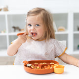 A toddler enjoys a meal using a spoon while wearing a Bumkins Junior Bib: Wander, eating from a colorful tray at a white table.