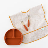 Bumkins Junior Bib: Wander lays flat on a white background with orange utensils on top of it, next to an orange divided plate.