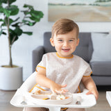 A toddler beams in a high chair while wearing a Bumkins Junior: Bib Wander, eating food from a beige divided plate.