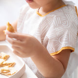 Close-up of a child wearing a Bumkins Junior Bib: Wander, holding a snack while more pieces fill a beige bowl on the white table.