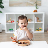 A toddler at a white table wears a Bumkins Junior Bib: Wander, smiling while eating from a divided plate, with toys on a white shelf behind.