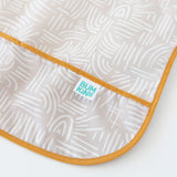 Close-up of Bumkins Junior Bib: Wander on a white background, featuring abstract white lines on beige, with focus on the catch-all pocket.