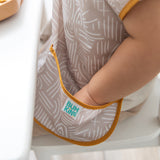 Close-up of a child at a white table, with their hand nestled in the catch-all pocket of a Bumkins Junior Bib: Wander.