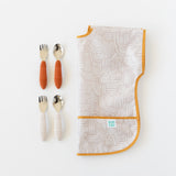 Two sets of baby utensils with orange & beige handles rest next to a folded Bumkins Junior: Bib Wander on a white background.