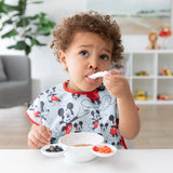 A young child in a Bumkins Junior Bib: Mickey Mouse Classic eats oatmeal, blueberries, and strawberries from a white bowl at a white table.