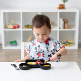 A toddler wearing a Bumkins Junior Bib: Mickey Mouse Classic eats with a fork at a white table, with toys and books in the background.