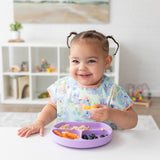 A girl in pigtails smiles, wearing a Bumkins Junior Bib 2 Pack: Stitch Star-ring Up Trouble as she eats from a purple plate at a white table.