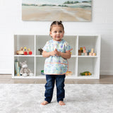A girl wearing a Bumkins Junior Bib 2 Pack: Stitch Star-ring Up Trouble stands in front of toy-filled shelves in a bright playroom.