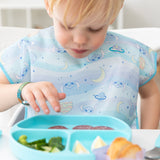 A toddler in a Bumkins Junior Bib 2 Pack: Stitch Star-ring Up Trouble looks down at a sectioned blue plate of food.