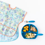 Bumkins Junior Bib 2 Pack: Stitch Star-ring Up Trouble lays stacked on a white background next to a blue divided plate with pasta, oranges, veggies, and utensils.