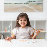 A child in a Bumkins Short-Sleeved Smock: Wander smiles, holding a paintbrush at a white table in a sunlit room.