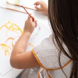 Close-up of a child wearing Bumkins Short-Sleeved Smock: Wander paints in orange and yellow at a white table.