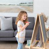 A young girl in a Bumkins Short-Sleeved Smock: Wander paints at an easel, surrounded by a cozy living room with a sofa and landscape art.