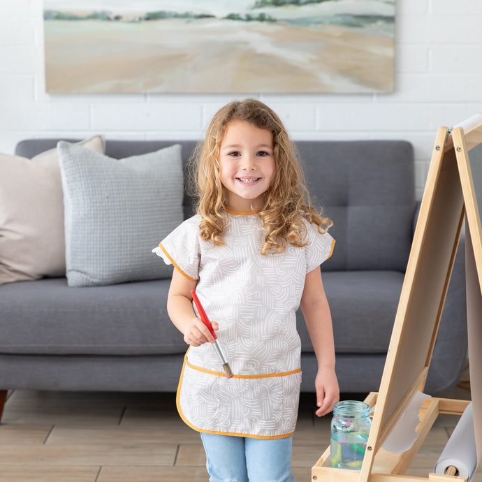 A young girl with curly hair wearing a Bumkins Short-Sleeved Smock: Wander stands in a living room, holding a paintbrush by a wooden easel.