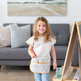 A young girl with curly hair wearing a Bumkins Short-Sleeved Smock: Wander stands in a living room, holding a paintbrush by a wooden easel.