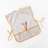 Bumkins Short-Sleeved Smock: Wander lays on a white background with two paintbrushes in the pocket.