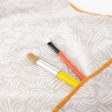 Close-up of two paintbrushes in the pocket of a Bumkins Short-Sleeved Smock: Wander.