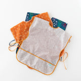 Bumkins Short-Sleeved Smock: Wander lays on top of a stack of similar art smocks on a white background.
