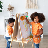 Two kids paint at an easel in a bright room; one smiles, wearing a Bumkins Short-Sleeved Smock: Grounded, while the other focuses.