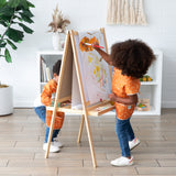 Two children, one in a Bumkins Short-Sleeved Smock: Grounded, paint freely on an easel in a bright room with shelves and a plant.