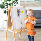 A child in a Bumkins Short-Sleeved Smock: Grounded holds a paintbrush by an easel with art, with a large plant and couch in the background.