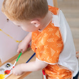 A child painting at an easel in a wearing a Bumkins Short-Sleeved Smock: Grounded, holding a brush.