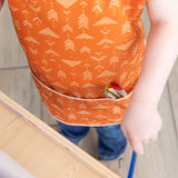 Close-up of a child in a Bumkins Short-Sleeved Smock: Grounded with paintbrushes in the pocket, painting at an easel.