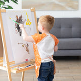 A toddler in a Bumkins Short-Sleeved Smock: Grounded paints at an easel with a paintbrush, with a couch in the background.